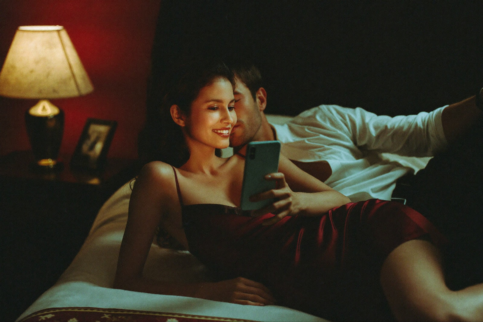 Um casal na cama, à luz do abajur, olhando juntos o celular